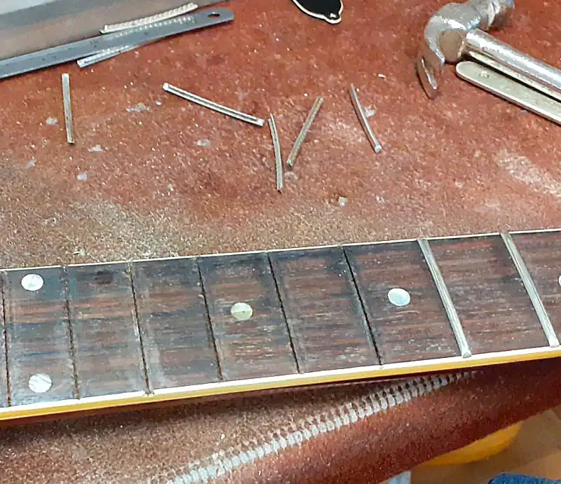 Guitar Fretwork and Refretting Close-up of a guitar rosewood fretboard undergoing a refret, showing removed fret wire pieces and luthier tools on a workbench.